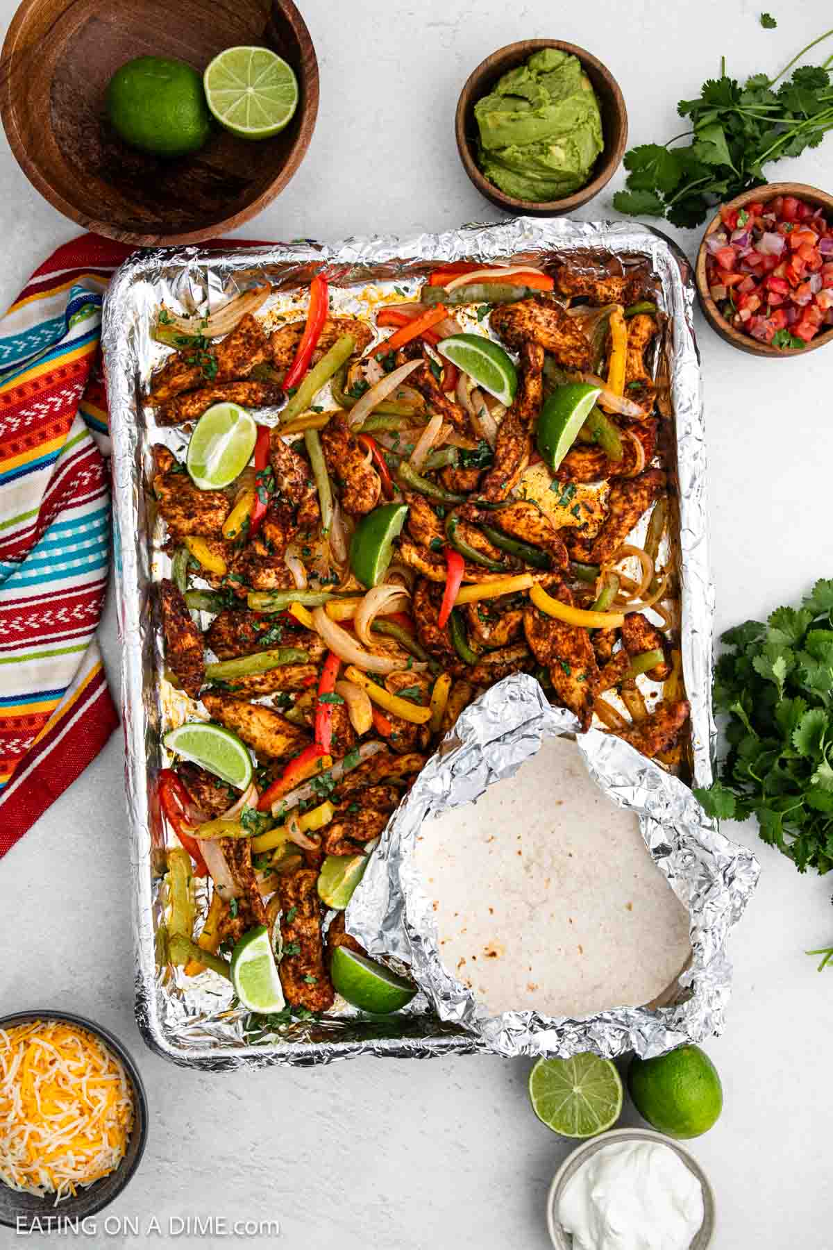 Seasoned Chicken Strips with sliced bell peppers and lime wedges, surrounded by foil-wrapped tortillas, bowls of guacamole, pico de gallo, cheese, sour cream, fresh cilantro, and a colorful napkin.