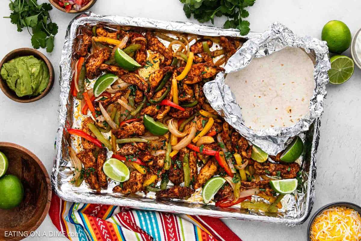 Sheet pan chicken fajitas with sliced peppers, onions, and lime wedges rest on a foil-lined sheet pan with foil packet of tortillas. Small bowl of guacamole, limes and shredded cheese surround the pan.