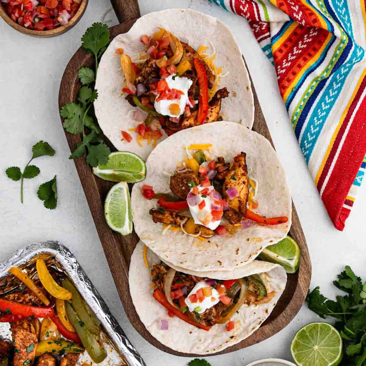 Three sheet pan chicken fajitas tacos with grilled peppers, onions, pico de gallo, and sour cream on a wooden board, garnished with lime wedges and cilantro. A colorful cloth napkin is beside the board.