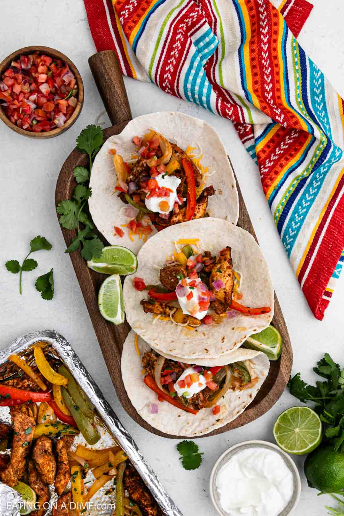 Three chicken fajitas with sautéed peppers and onions are served on tortillas, topped with sour cream and pico de gallo, on a wooden board with lime wedges. A colorful napkin, cilantro, and a tray of sheet pan chicken fajitas ingredients are nearby.