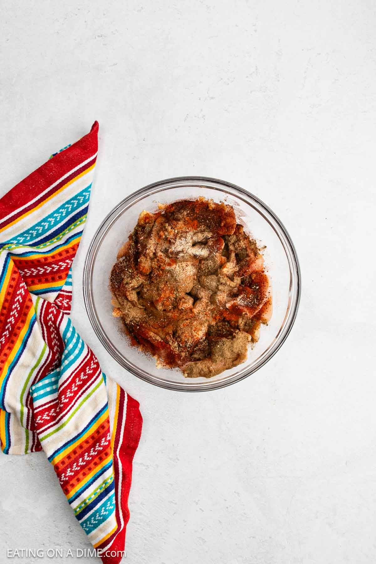 A glass bowl filled with seasoned raw chicken pieces, perfect for sheet pan chicken fajitas, sits on a white surface next to a colorful striped kitchen towel.