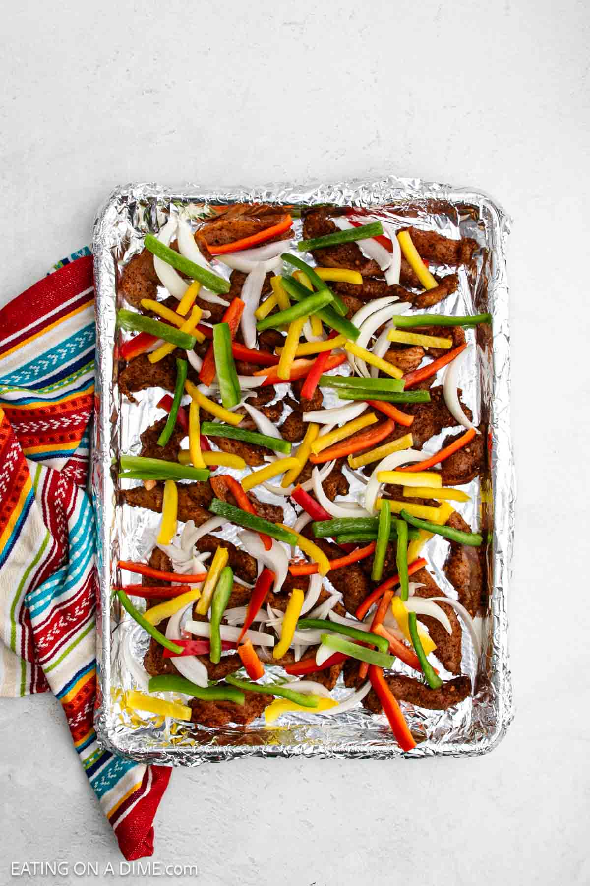 A foil-lined baking sheet topped with seasoned sheet pan chicken fajitas, featuring sliced bell peppers and onion strips, sits next to a colorful striped cloth on a light surface.