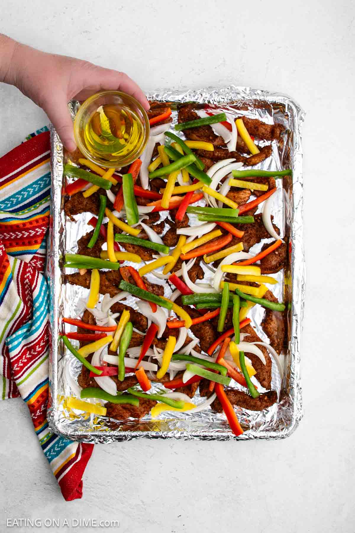 A hand holding a glass of yellow drink over a foil-lined baking sheet with seasoned sheet pan chicken fajitas—chicken strips, sliced red, yellow, and green bell peppers, and onions—next to a colorful striped towel.
