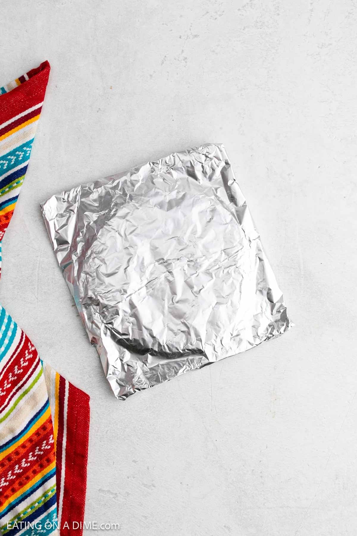 A round item wrapped in aluminum foil sits on a light gray surface next to a colorful striped cloth, evoking the vibrant look of sheet pan chicken fajitas with its red, blue, yellow, and green patterns.