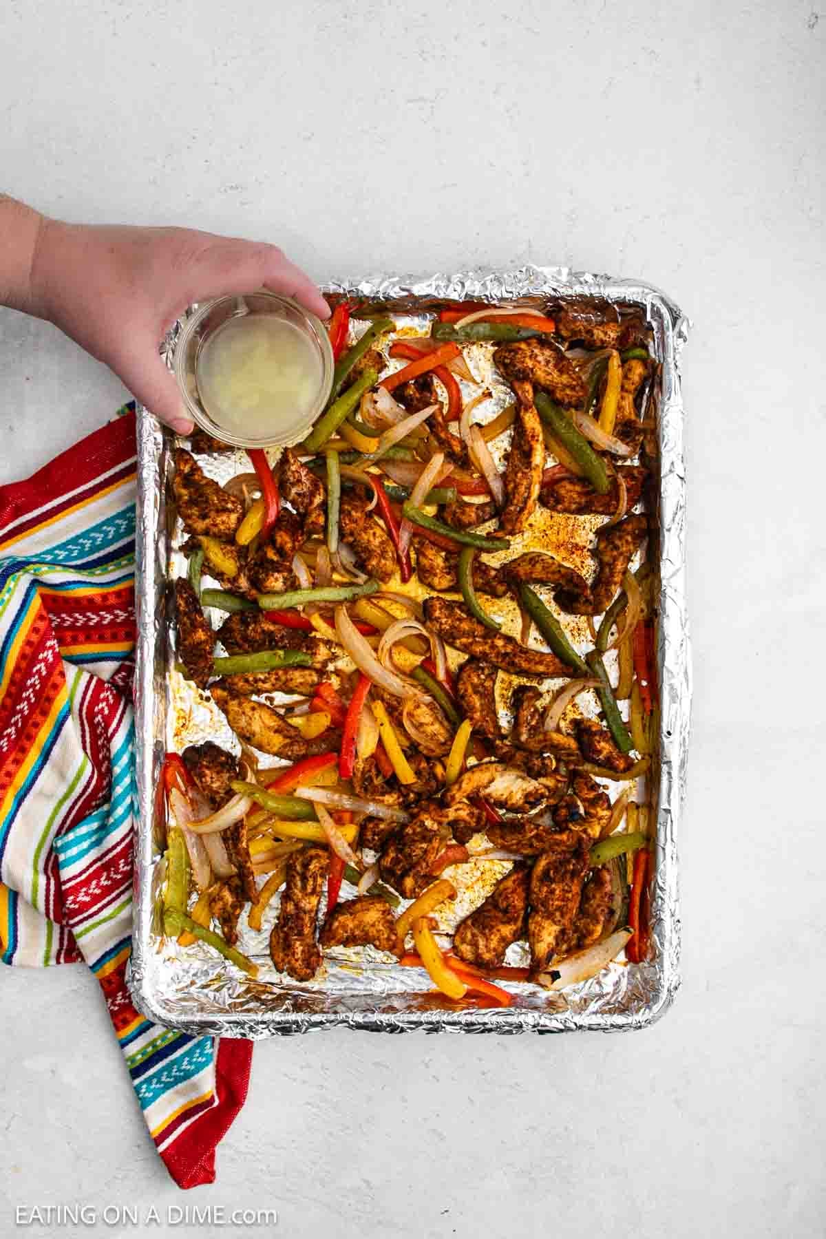 A foil-lined baking sheet filled with sheet pan chicken fajitas, sliced bell peppers, and onions. A hand holds a drink above the pan, which rests on a colorful striped cloth.