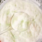 A ladle scooping frothy, white foam from a bowl of light green, creamy Cherry limeade Sherbet Punch. The bubbly punch appears airy and refreshing.