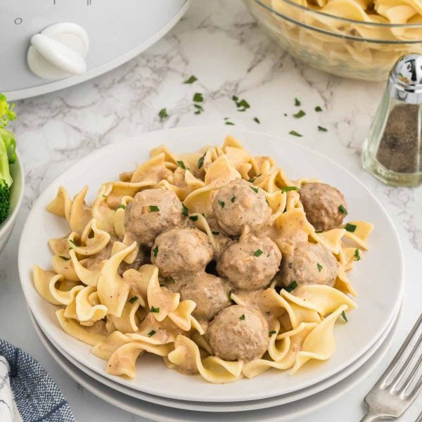 A plate of egg noodles topped with creamy slow cooker Swedish meatballs and garnished with chopped parsley, set on a marble table with a bowl of extra noodles, a salt shaker, and broccoli nearby.