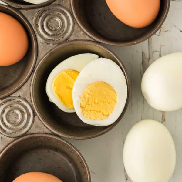 A close-up of Boiled Eggs in the Oven nestled in a metal muffin tin, with a halved hard-boiled egg revealing its yellow yolk and peeled eggs resting on a white wooden surface nearby.