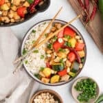 A bowl of kung pao chicken recipe with stir-fried chicken, zucchini, bell peppers, peanuts, and dried chili peppers served over white rice and topped with green onions. Chopsticks rest on the bowl; fresh ingredients are scattered nearby.