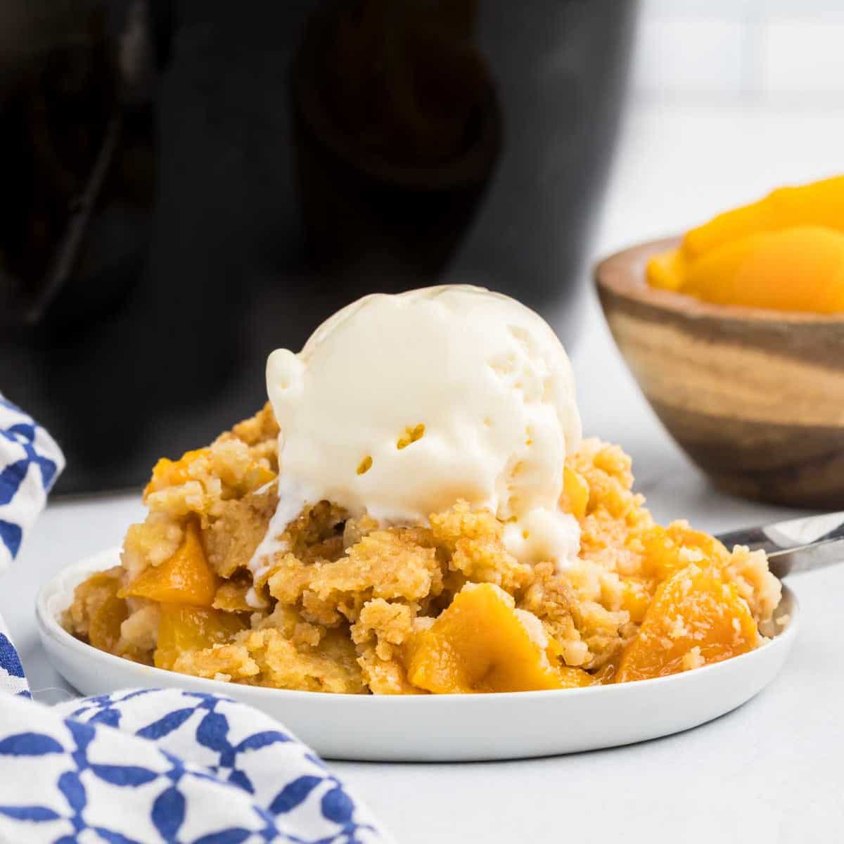 A serving of Crock Pot Peach Dump Cake topped with vanilla ice cream sits on a white plate, with a bowl of sliced peaches and a blue and white napkin in the background.