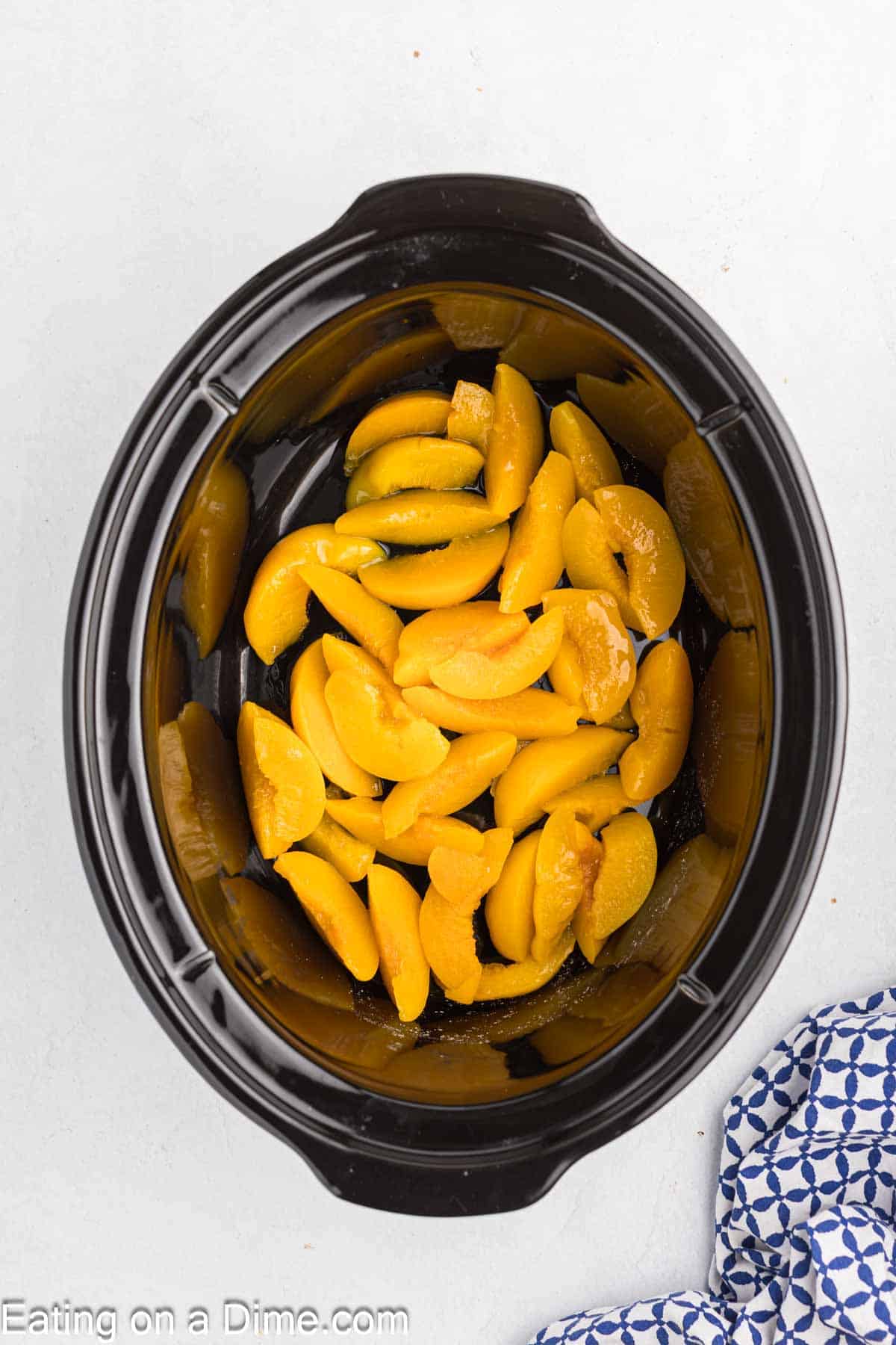 Sliced peaches in syrup are placed in a black slow cooker on a white surface, ready to be transformed into an easy Crock Pot Peach Dump Cake; part of a blue-and-white patterned cloth is visible in the corner.