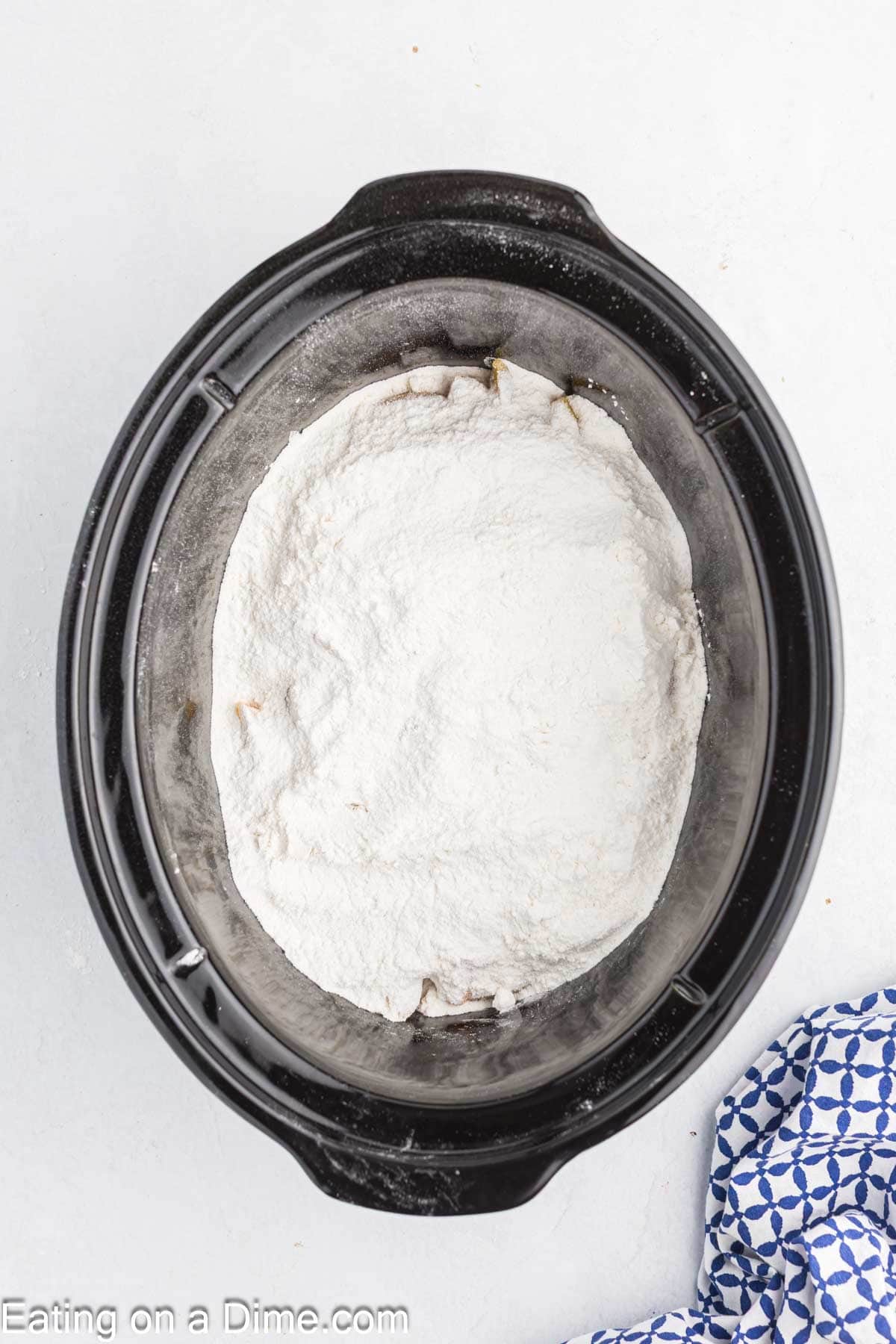 A Crock Pot Peach Dump Cake is prepared in a slow cooker filled with a layer of white flour or powdered baking mix, all set on a white surface beside a blue and white patterned cloth.