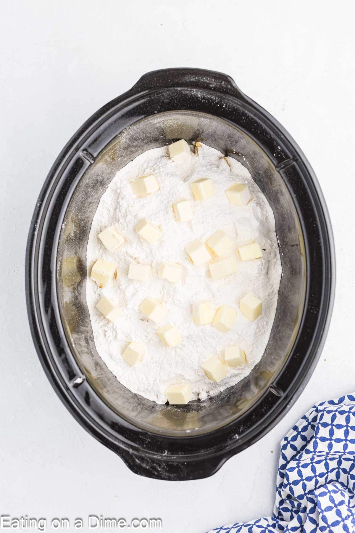 A Crock Pot Peach Dump Cake sits in a slow cooker, with a layer of white cake mix topped by evenly distributed cubes of butter, while part of a blue-patterned cloth is visible in the corner.