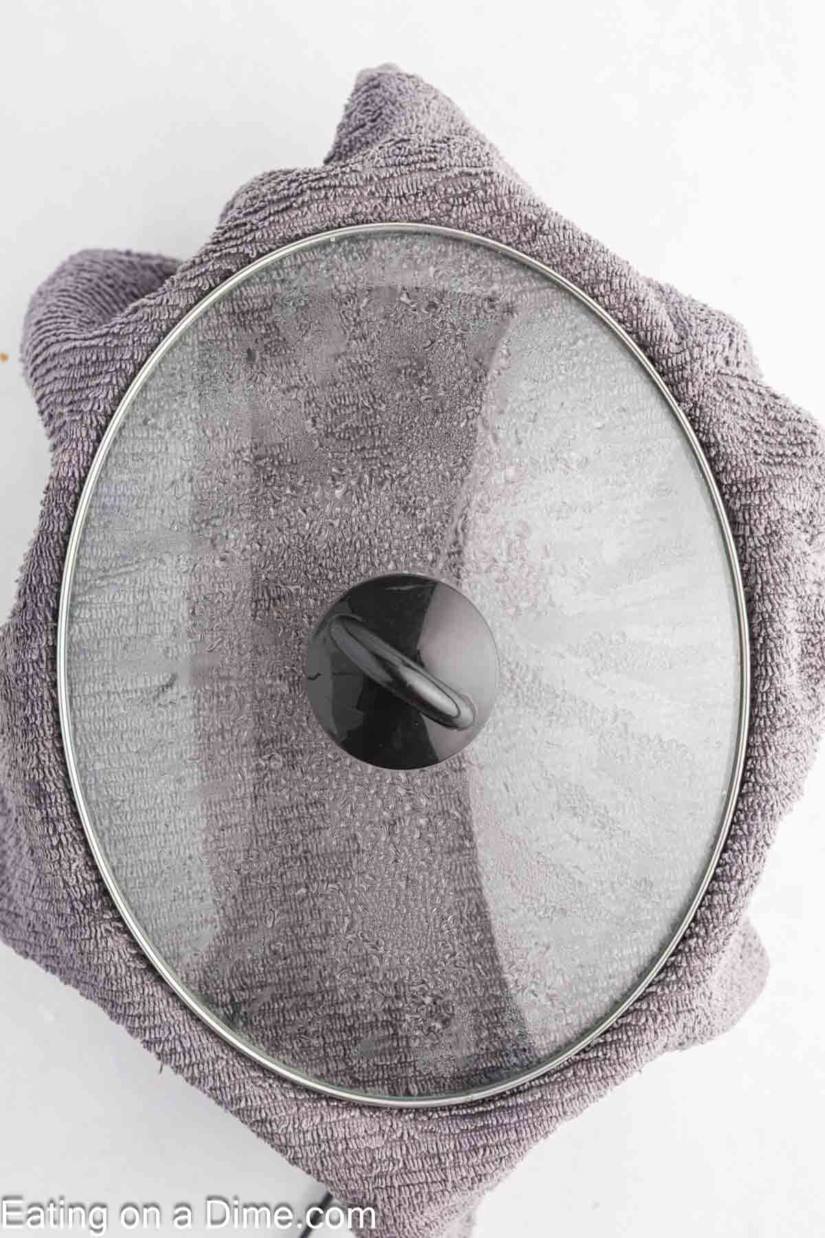 A glass pot lid sits on top of a gray towel, viewed from above. The towel covers a surface, and part of the website name "Eating on a Dime.com" is visible in the corner—perfect for preparing Crock Pot Peach Dump Cake.