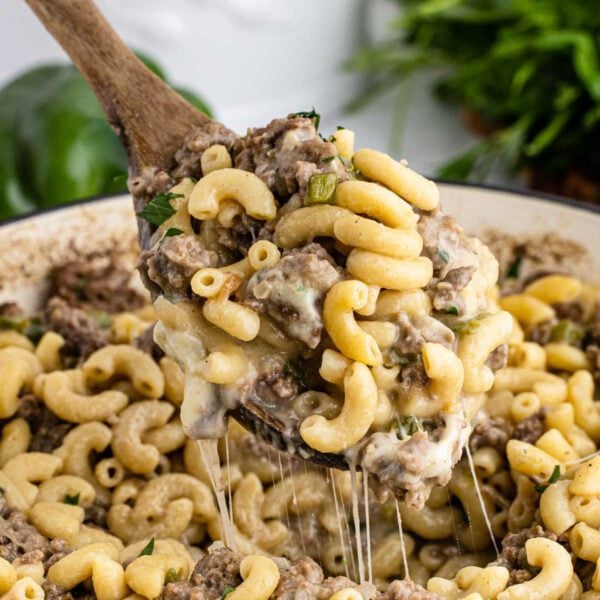 A wooden spoon lifts a serving of cheesy ground beef and macaroni from a pot, with stretchy melted cheese visible among the pasta and beef mixture—just like in a classic Philly cheesesteak pasta recipe. Fresh herbs are sprinkled on top.