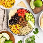 A delicious taco bowl recipe featuring rice, ground meat, black beans, corn, diced tomatoes, avocado, tortilla chips, and a lime wedge, surrounded by fresh lime slices, corn, and cilantro on a light surface.