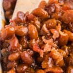 Close-up of a Baked Beans Recipe in a rich, glossy sauce with pieces of bacon, being stirred with a wooden spoon.