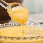 A spoon lifts creamy yellow caesar salad dressing sauce from a decorative glass bowl, with the sauce dripping smoothly back into the dish.