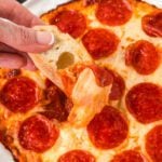 A hand holding bread scoops up gooey cheese and pepperoni, stretching from a delicious baked Pepperoni Pizza Dip—melty, savory perfection in every bite.