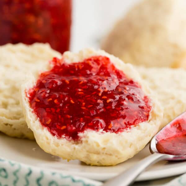 A biscuit cut in half, topped with bright red Raspberry Freezer Jam, sits on a plate. A spoon with some jam rests nearby. Another biscuit and a jar of jam are blurred in the background.
