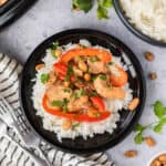A black plate of white rice topped with Slow Cooker Thai Peanut Chicken, red bell peppers, peanuts, and fresh cilantro, with extra rice and cilantro leaves nearby on a gray surface.