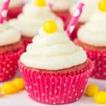 A pink strawberry lemonade cupcake with white frosting, topped with a yellow candy and a pink polka-dot straw, sits in a pink polka-dot wrapper. More cupcakes and yellow candies are in the background.