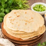 A stack of homemade flatbreads made with a classic flour tortilla recipe rests on a wooden plate, surrounded by fresh cilantro and small bowls of chopped green onions and other ingredients on a wooden table.