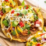 A close-up of a crispy tostada topped with refried beans, shredded chicken, cheese, diced avocado, tomato, red onion, and fresh cilantro. This colorful Chicken Tostadas Recipe is served on parchment paper for a vibrant meal.