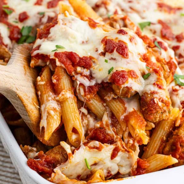 A wooden spoon scoops easy chicken parmesan pasta—baked penne topped with melted cheese, tomato sauce, and herbs—from a casserole dish.