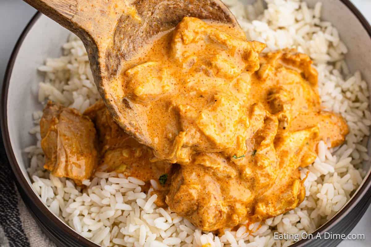 Close up image of butter chicken over white rice in a bowl with a wooden spoon