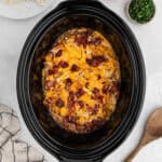 A Crock Pot Chicken Bacon Ranch Recipe features a slow cooker filled with shredded chicken, melted cheese, and crispy bacon bits, surrounded by a small bowl of chopped herbs, a wooden spoon, and a striped towel.