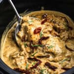 Creamy Crock Pot Sun Dried Tomato Chicken is covered in a rich herb sauce with rosemary and sun-dried tomatoes, served hot from the slow cooker with a large spoon.