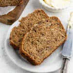Two slices of Healthy Banana Bread Recipe sit on a white plate, with a butter knife and a dish of butter nearby. A wooden board with more bread is in the background.