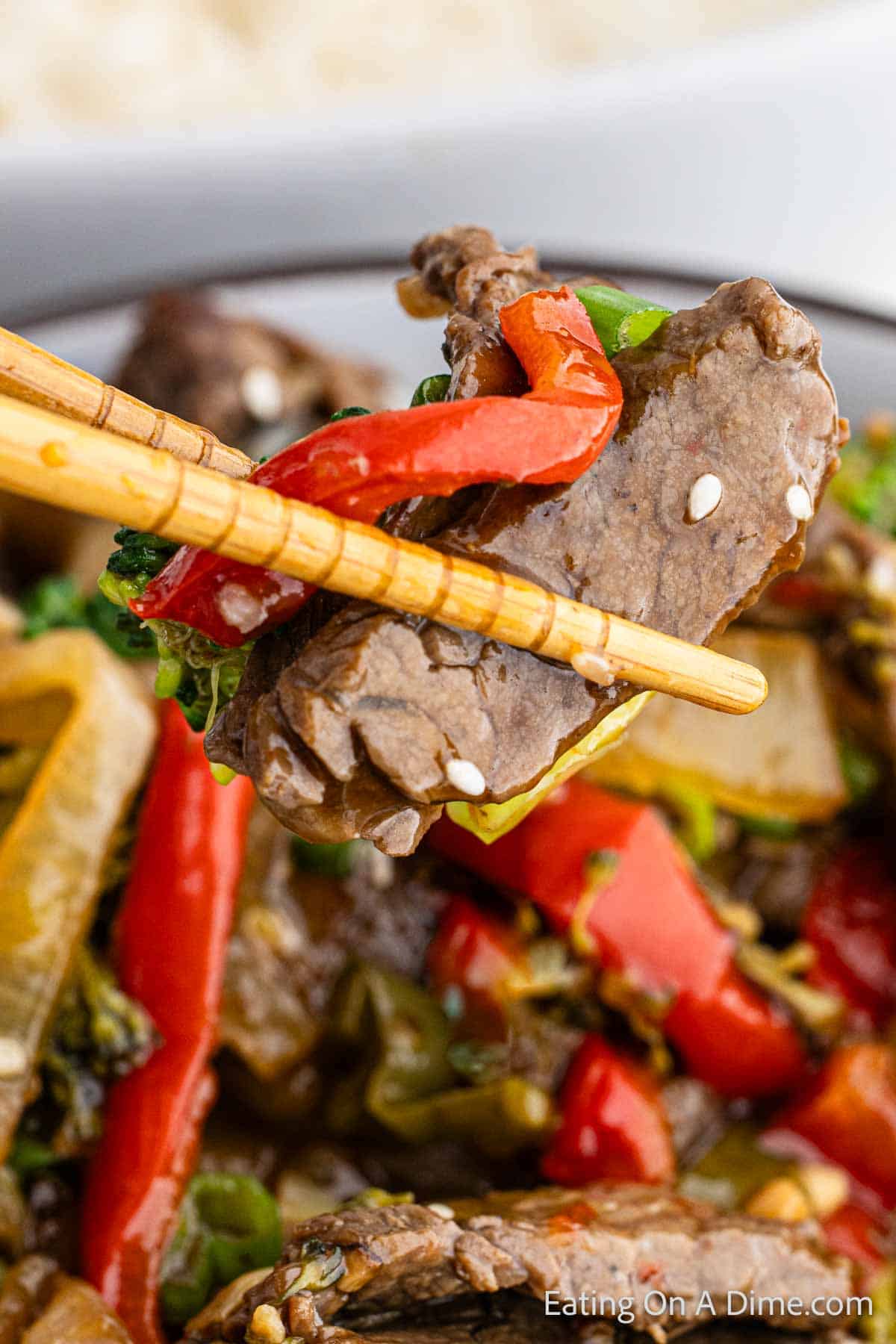 Chopsticks with a bite of beef and red peppers with a bowl of beef stir fry in the background