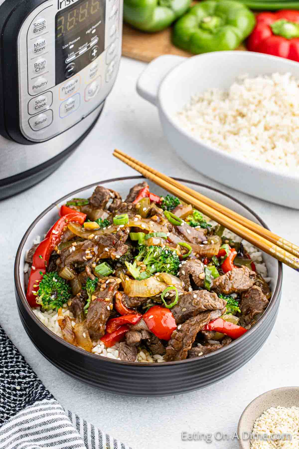 Beef Stir Fry in a bowl with broccoli, red peppers and onions. Chopsticks are on top of the bowl with a bowl of white rice in the background