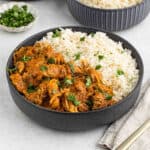 A black bowl filled with white rice and shredded Instant Pot Chicken Tikka Masala in a vibrant red curry sauce, garnished with fresh cilantro. A small dish of chopped herbs and another bowl of rice are in the background.