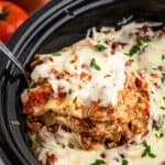 A serving spoon lifts a portion of cheesy slow cooker sausage lasagna, revealing layers of pasta, savory sausage meat sauce, and melted cheese, garnished with chopped parsley.