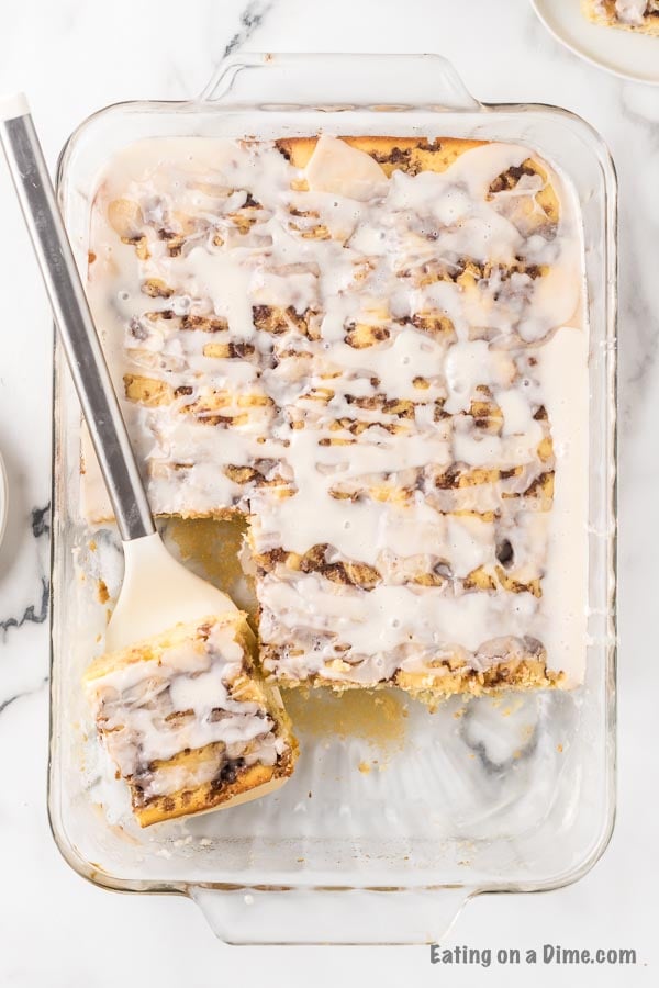 Overview of the cinnamon roll cake in a glass pan with the glaze drizzled on top of it with a piece being served out of it. 