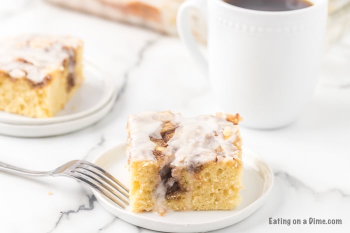 A piece of the cinnamon roll cake on a white plate with a fork with another piece on a plate in the back ground.