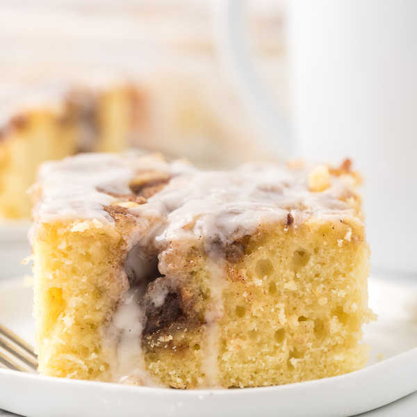 A close up of the cinnamon roll cake on a white plate with a cup of coffee behind it. 