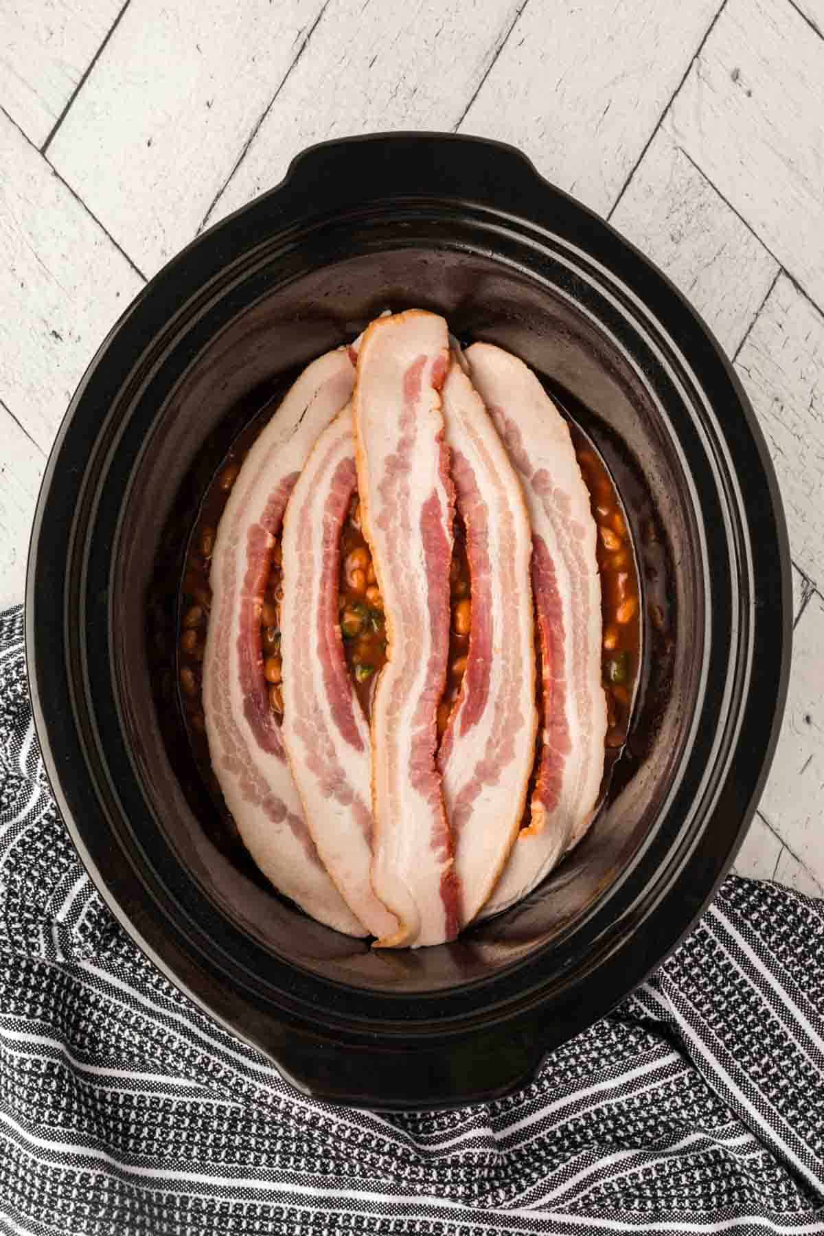 Placing the pork and beans mixture into the slow cooker and topping with strips of bacon