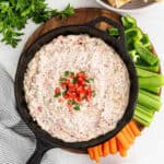 A cast iron skillet filled with sausage cream cheese dip, topped with diced tomatoes and green onions, surrounded by celery sticks, carrot sticks, green bell pepper slices, and fresh parsley on a wooden board.