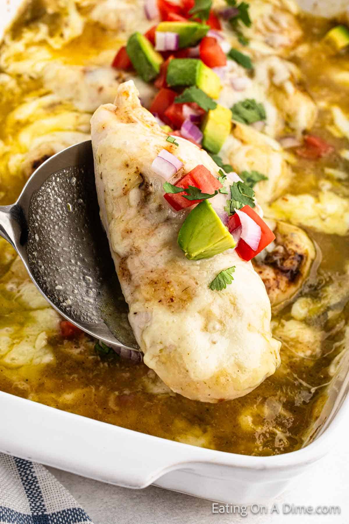 Baked chicken enchilada in a baking dish in the background with a serving on a spoon topped with avocado, tomato and fresh cilantro