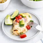 A plate of Baked Enchilada Chicken breast topped with melted cheese, diced avocado, tomato, red onion, and cilantro, served with lime wedges. A fork rests on the plate and a bowl of salad is visible in the background.