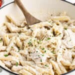Creamy chicken penne pasta with tender pieces of chicken, topped with shredded parmesan cheese and chopped herbs, served in a white pot with a wooden spoon.