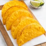 Five homemade taco shells are arranged upright in a row on a white napkin atop a wooden board. In the background, lime wedges are visible on a small dish.