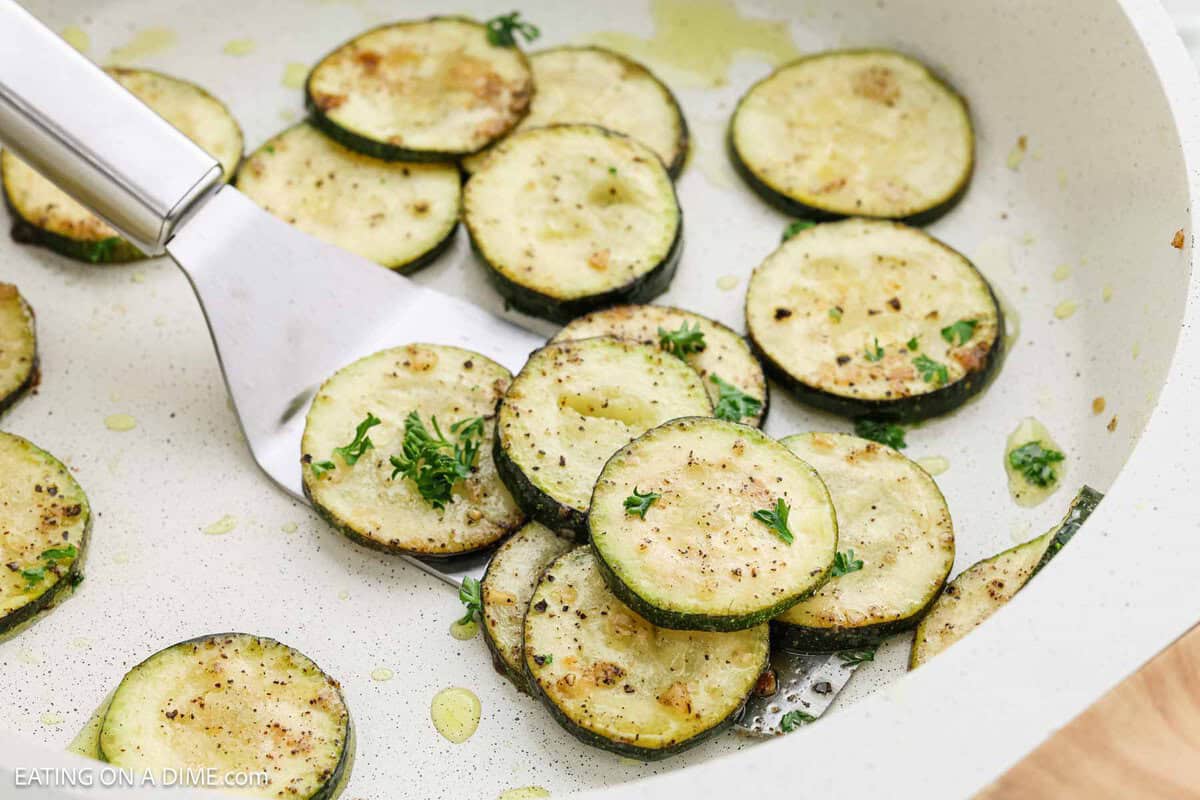 Pan-fried zucchini slices sizzle in the pan, perfectly seasoned with herbs and spices. A spatula lifts the golden bites as chopped parsley adds a fresh garnish. Cooked to perfection in olive oil, this dish is a delightful medley of flavors.