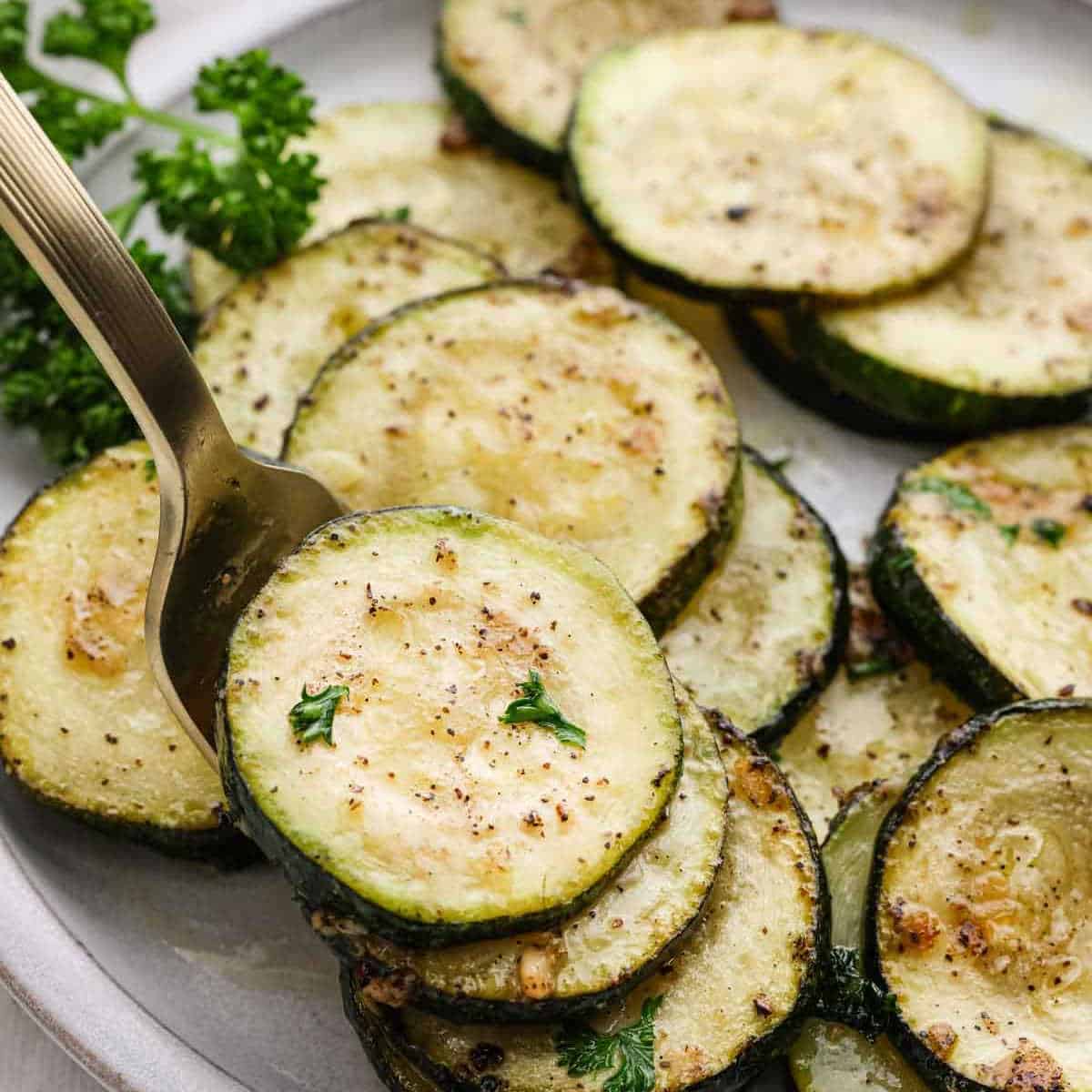 A close-up of pan-fried zucchini slices on a plate, seasoned with aromatic herbs and spices. Visible black pepper specks and bits of parsley enhance the delicious visual. A fork holds one slice, amplifying the appetizing freshness of the presentation.