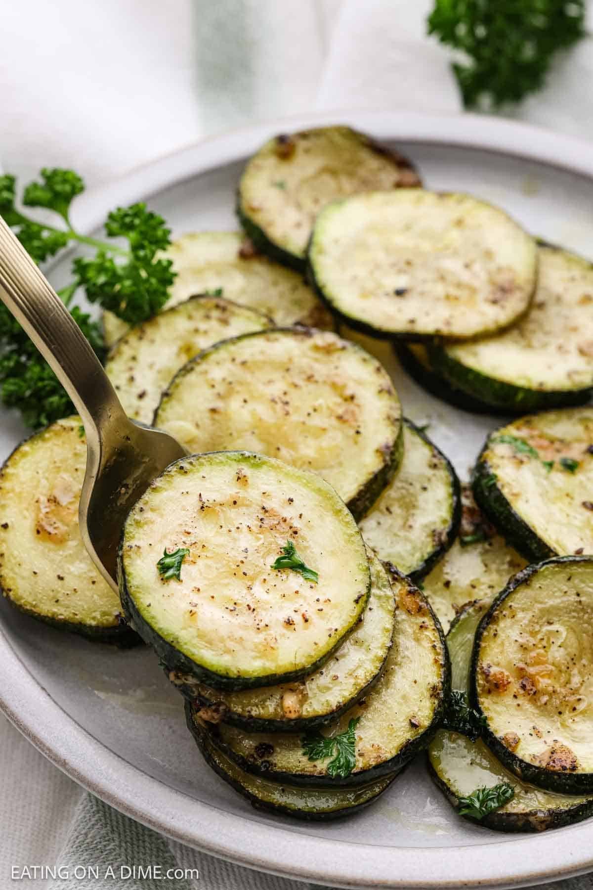 A white plate features pan-fried zucchini slices, artfully garnished with parsley. A fork is gracefully lifting one perfectly seasoned slice, its crispy edges revealing the medley of herbs and spices that enhance its flavor.