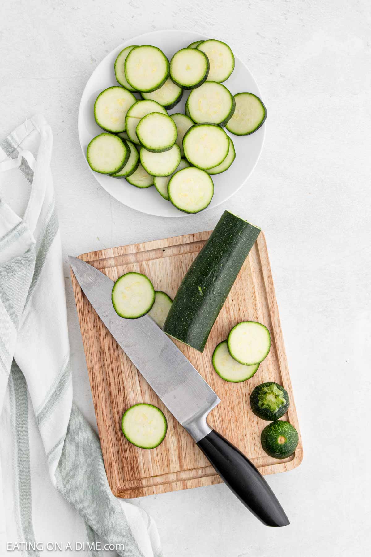 A wooden cutting board holds a partially sliced zucchini and a knife, perfect for prepping pan fried zucchini. Nearby, a white plate displays more slices, ready for the pan. A kitchen towel is draped beside the board on the light gray surface.